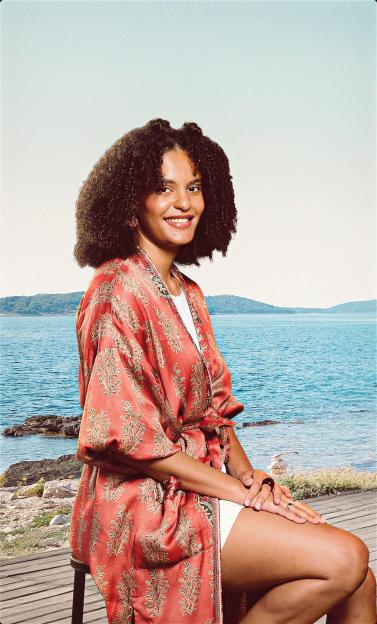 A smiling woman in a patterned robe sits on a stool by the sea.