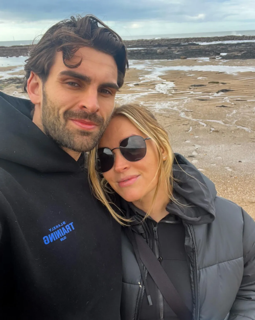 A man in a black hoodie and a woman in sunglasses and a black puffer jacket stand on a sandy beach.