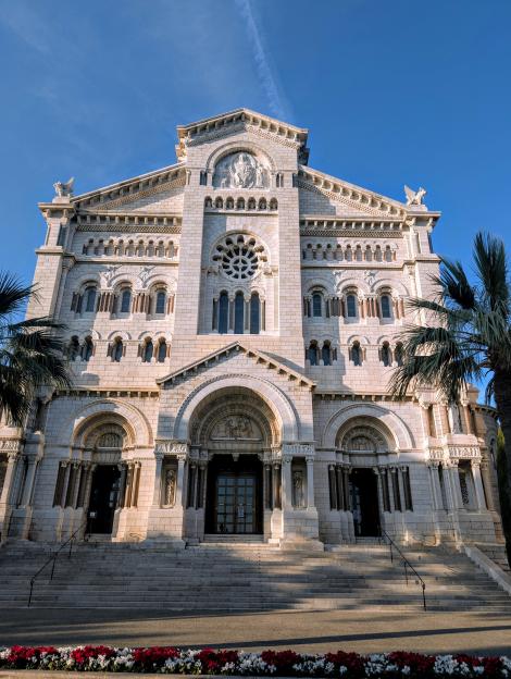 The Monaco Cathedral, a large white stone building, with multiple arched doorways and windows, and palm trees framing it, against a clear blue sky.