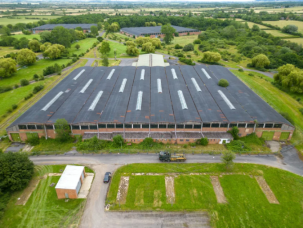 Aerial view of a large, long industrial building with a dark roof and many skylights, surrounded by green fields and scattered trees.