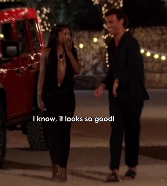 Two people, one woman and one man, walking outside at night next to a red car, the woman covering her mouth with her hand, both smiling.