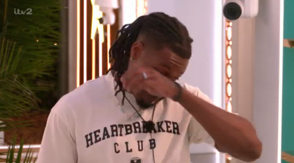 Man with dreadlocks wearing a white "Heartbreaker Club" shirt, crying with his hand covering his eyes.