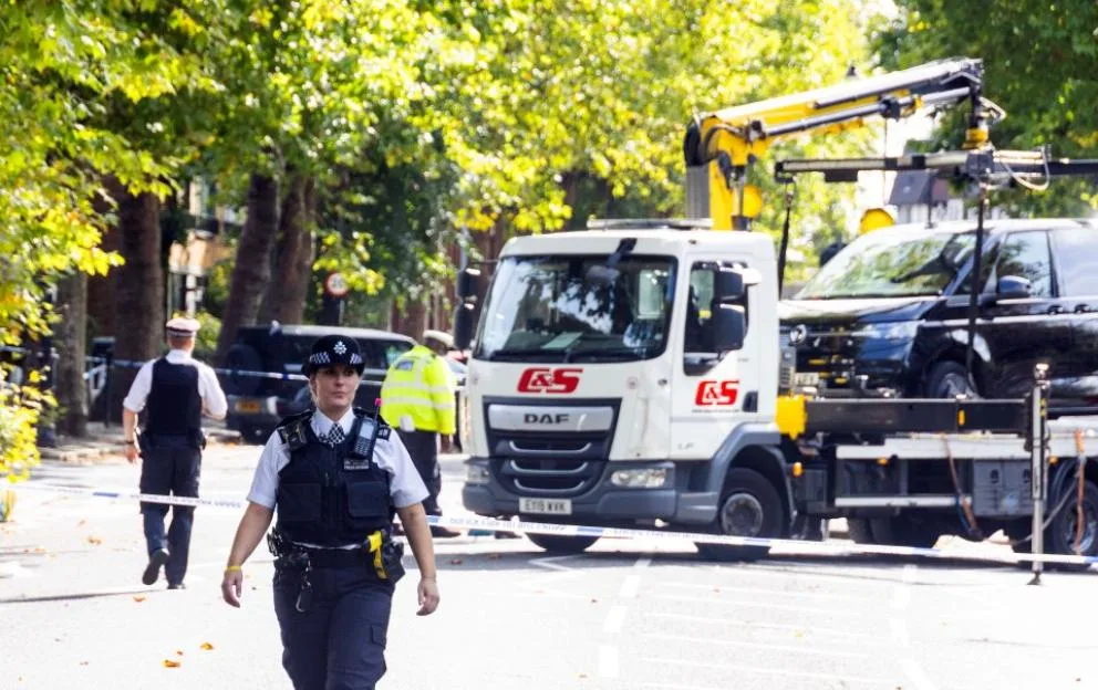 A police officer standing in a street cordoned off with blue and white tape, with a tow truck and another police officer in the background.