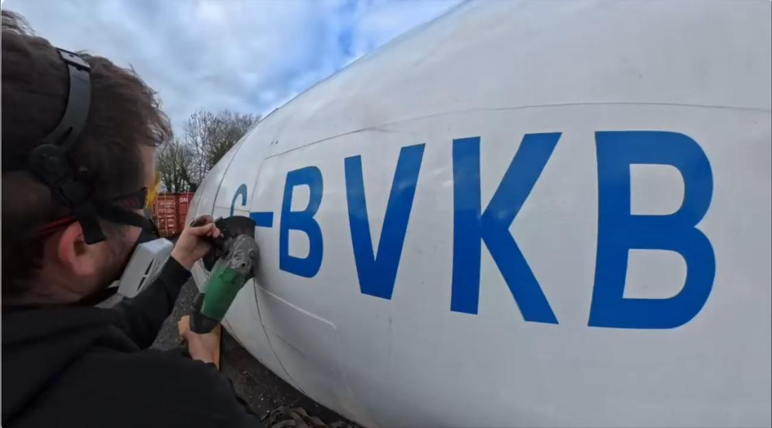 A man using an angle grinder to remove letters from the side of a white airplane fuselage.
