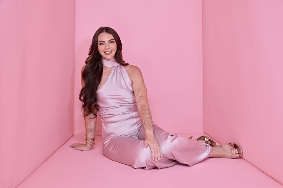 Amy Salmon in a full-body shot, smiling, wearing a pink satin halter top and pants, seated on a pink floor.