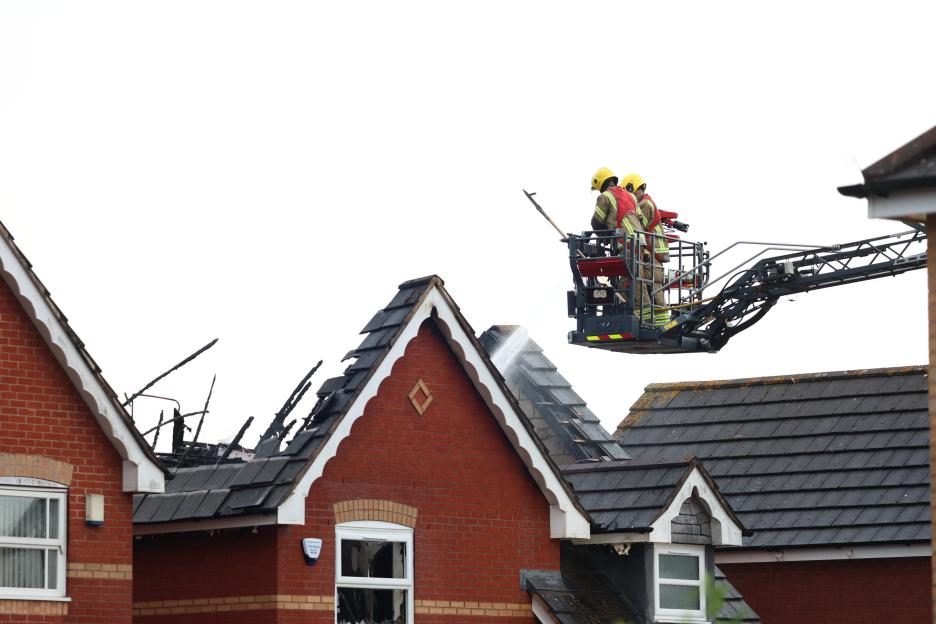 Firefighters on an elevated platform hose down the burned roof of a brick house.