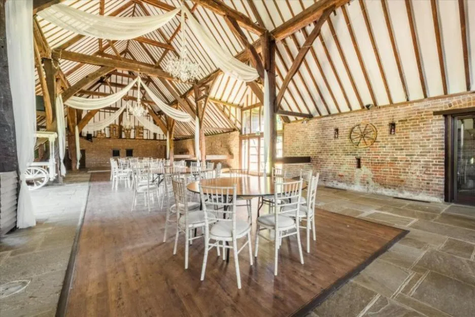 Interior of a spacious rustic barn with a high vaulted wooden ceiling, exposed beams, brick walls, and white draped fabric, set up with round tables and white chairs for an event.