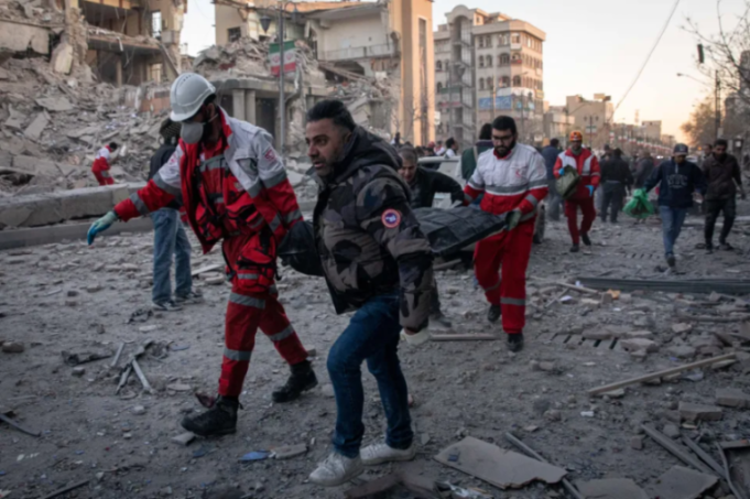Rescuers carrying a body through rubble in a damaged city.