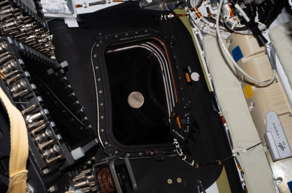 The Moon seen through a spacecraft's window, surrounded by wires and equipment.