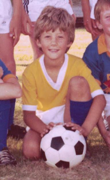 Chris Jones as a child in a yellow soccer uniform kneeling on grass with a soccer ball.