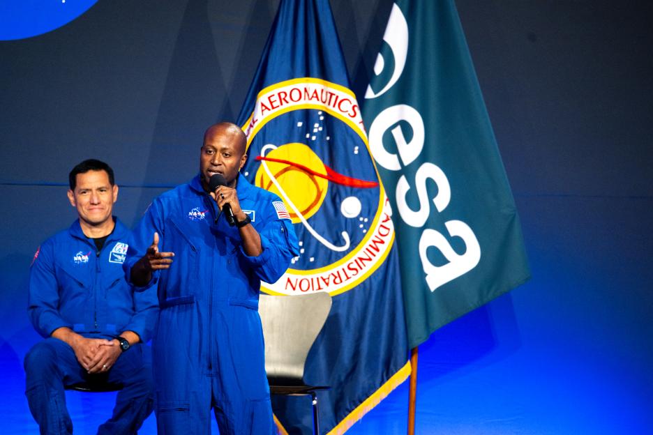 Two men in blue flight suits stand before a NASA flag, one speaking into a microphone.