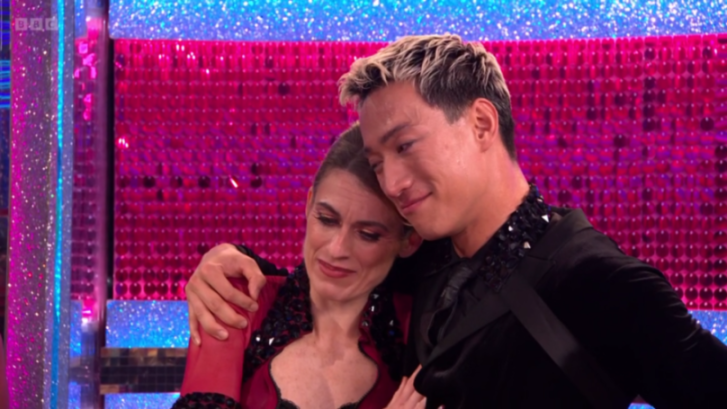 A blonde man wearing a black suit with studded shoulders hugs a woman in a red top with a black lace trim.