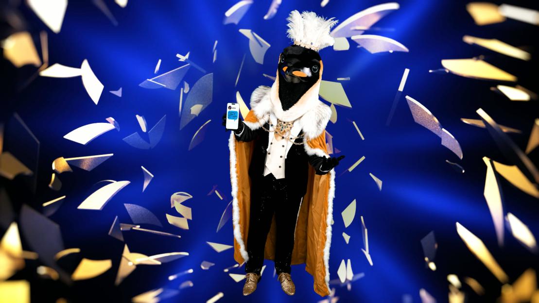 The Penguin from The Masked Singer in a costume featuring an orange cape, holding a phone.