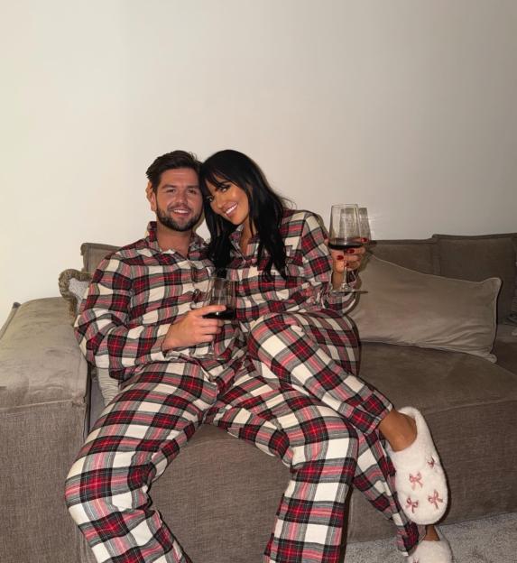 A couple in matching red, white, and black plaid pajamas on a couch, each holding a glass of red wine.