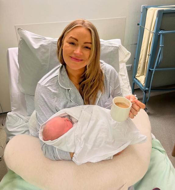 Woman holding newborn baby in hospital bed, drinking tea.