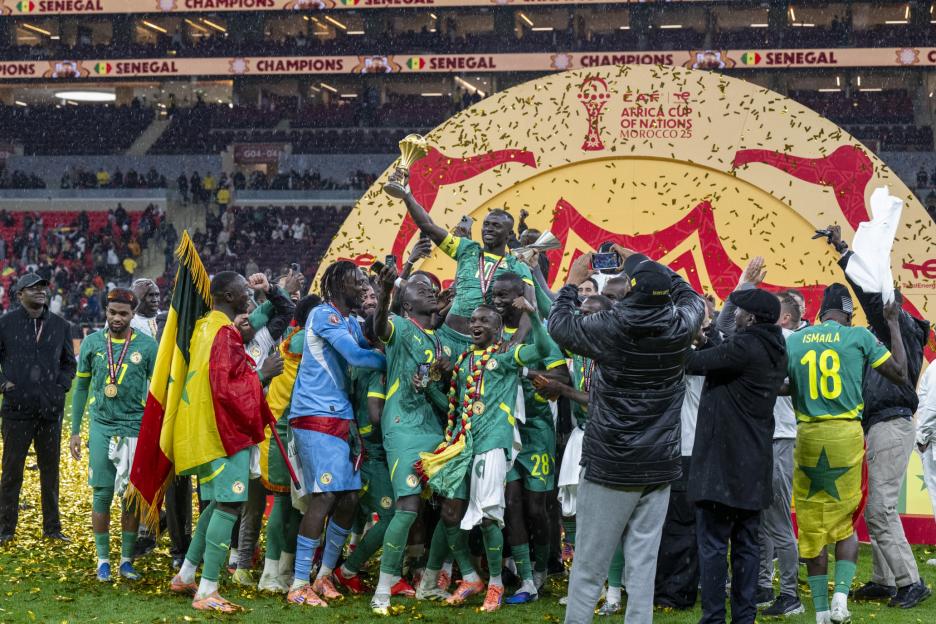 Senegal team celebrates winning the AFCON title, with one player holding the trophy aloft under falling confetti.