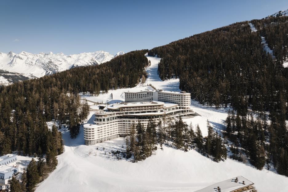 Aerial view of a large, modern white resort hotel built into a snowy mountain ski slope, surrounded by pine trees with snow-capped mountains in the background.
