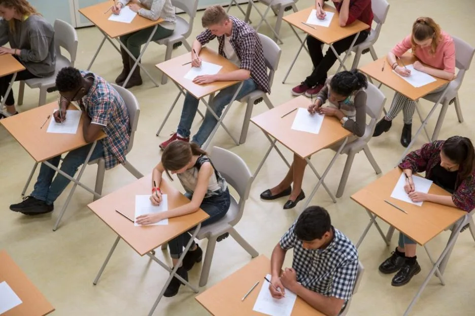 An elevated view shows a diverse group of students sitting at individual desks and writing GCSE exams.