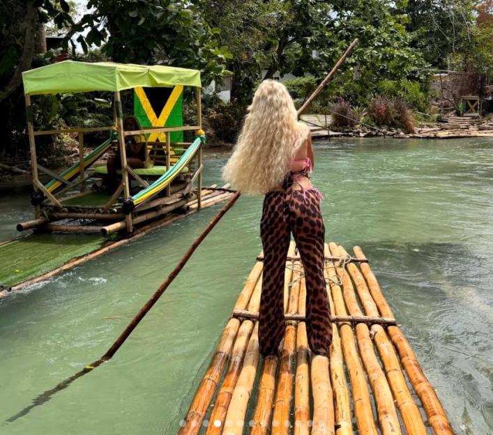 A person with long blonde hair and leopard-print pants standing on a bamboo raft, looking towards another raft with a Jamaican flag.