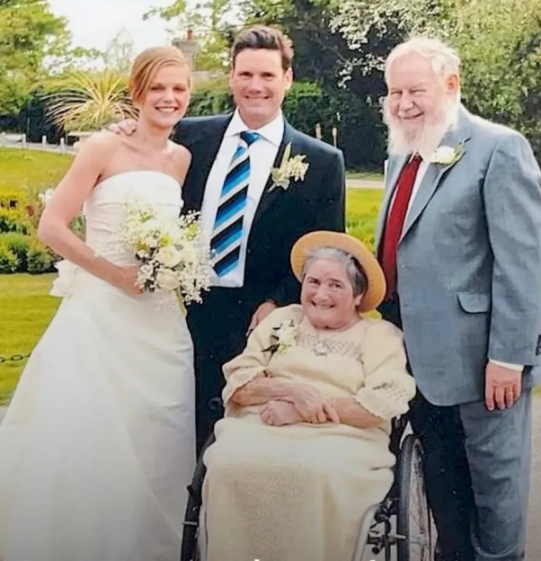 A bride and groom pose with two older relatives, one in a wheelchair, all smiling.