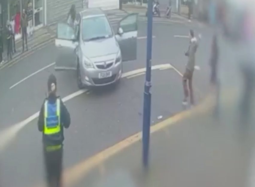 A car with open doors on the street, with people standing around and a person in a safety vest walking away.