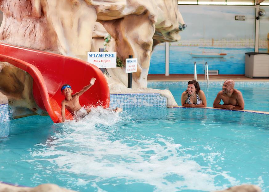 A child exiting a red water slide in an indoor pool as two adults watch and laugh.