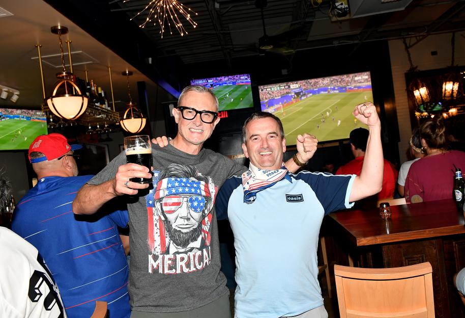 Two men pose with drinks in a pub, with a soccer match playing on screens in the background.