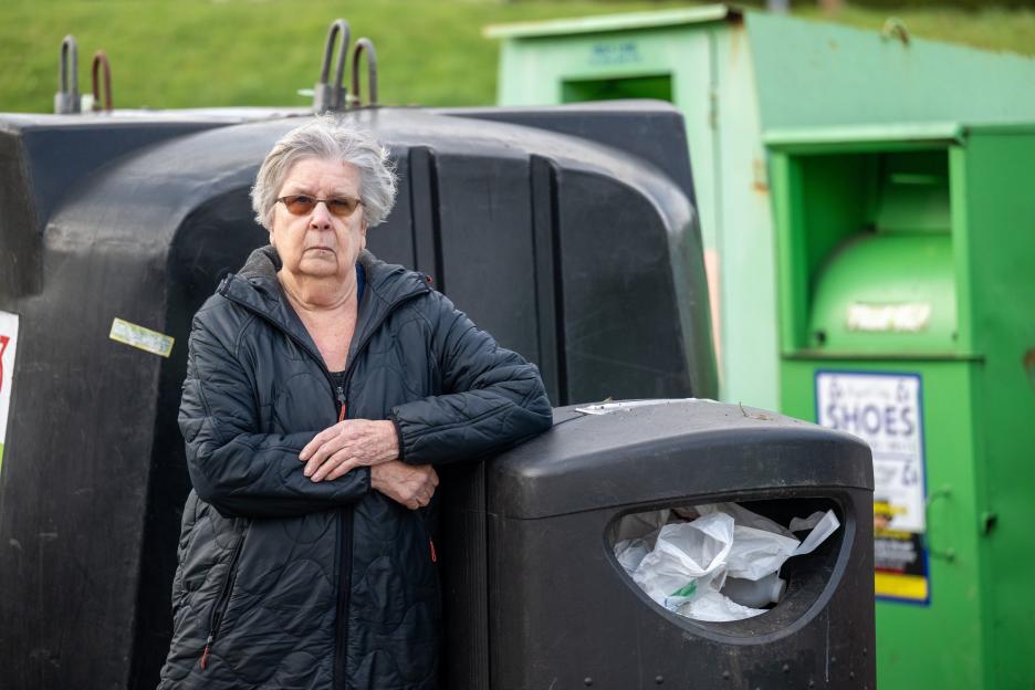 An elderly woman in a dark jacket and sunglasses stands next to a black recycling bin, with large green recycling bins in the background.