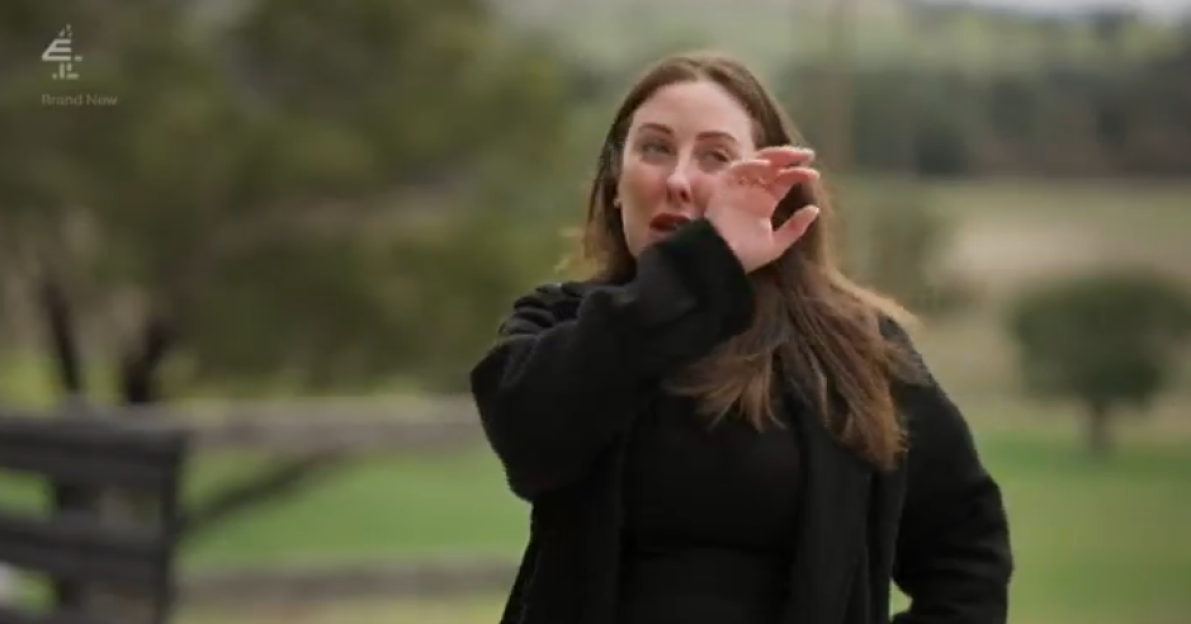 A woman with long brown hair, wearing a black top and coat, wiping her eye with her hand.