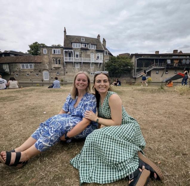 Two young women sitting on dry grass by a river, with historic buildings and people in the background.