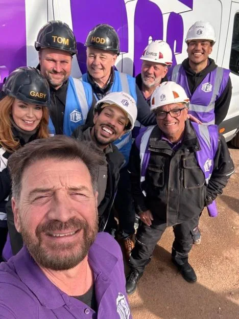 A group of eight people, including Nick Knowles taking the selfie, wearing high-visibility vests and hard hats with their names on them, posing in front of a van.