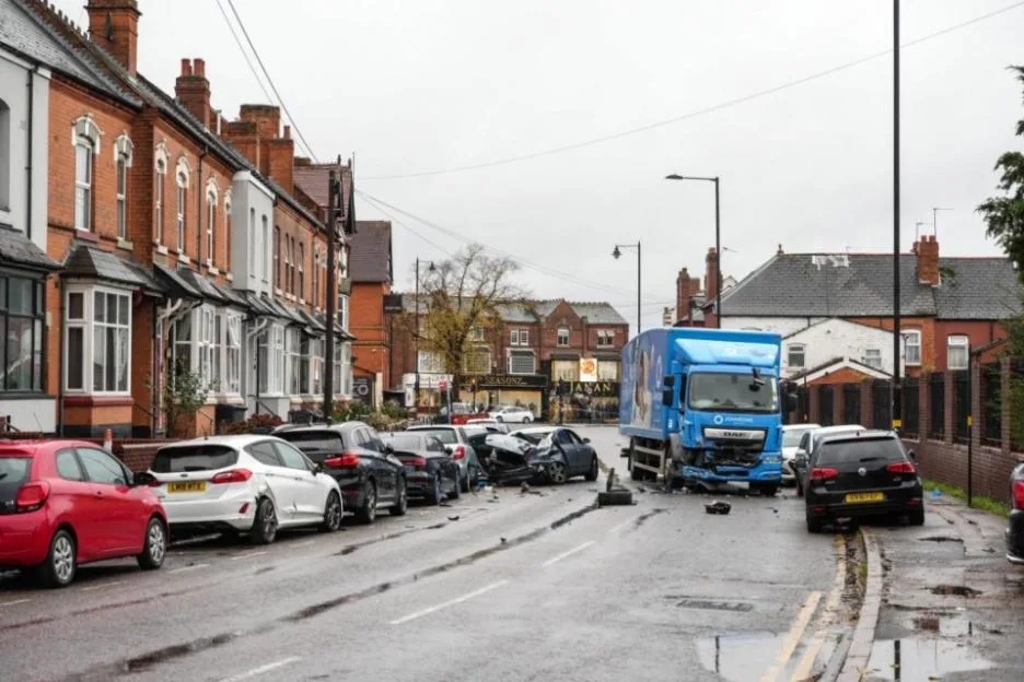 A street scene showing a blue lorry and several badly damaged parked cars after a collision.