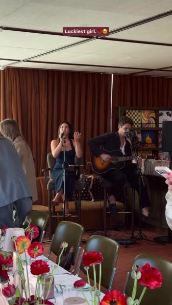 A woman sings into a microphone while a man plays acoustic guitar in a restaurant or event space.