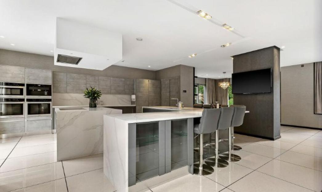 A spacious, modern kitchen with light gray and white tones, featuring a large island with gray bar stools, built-in ovens, and a flat-screen TV mounted on a dark pillar.