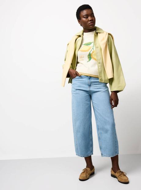 A woman wearing a white sweater with a lemon graphic and the words "DOLCE VITA," a light green jacket, light-wash wide-leg jeans, and brown boat shoes.
