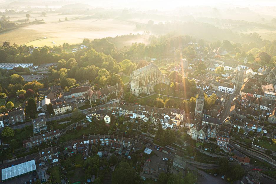 Aerial view of Malmesbury, Wiltshire, with the abbey in the center, surrounded by houses and fields, seen on a misty morning with sunlight streaming through.
