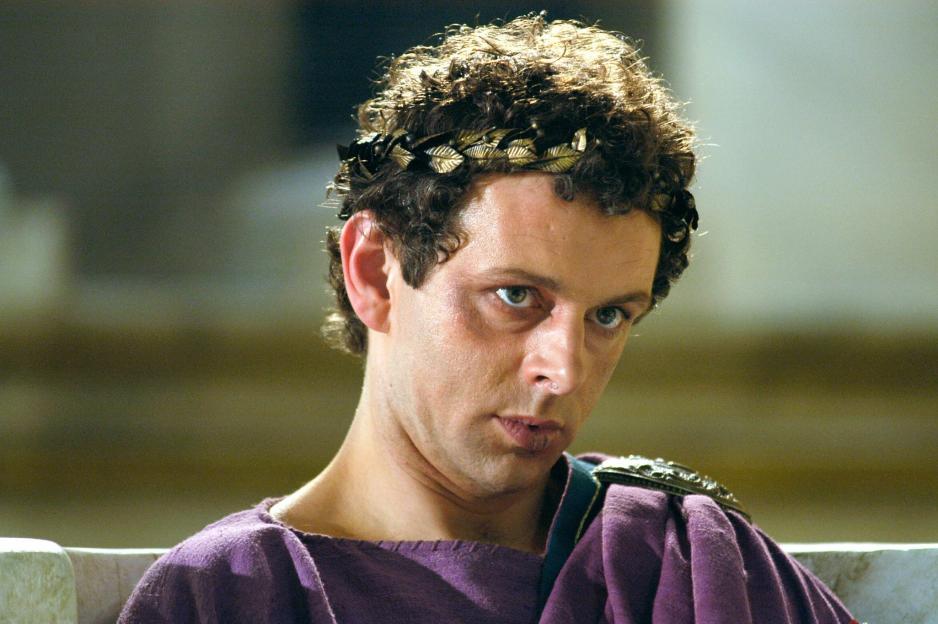 A man with curly brown hair, wearing a gold laurel wreath, purple toga, and green tunic, looks towards the viewer.