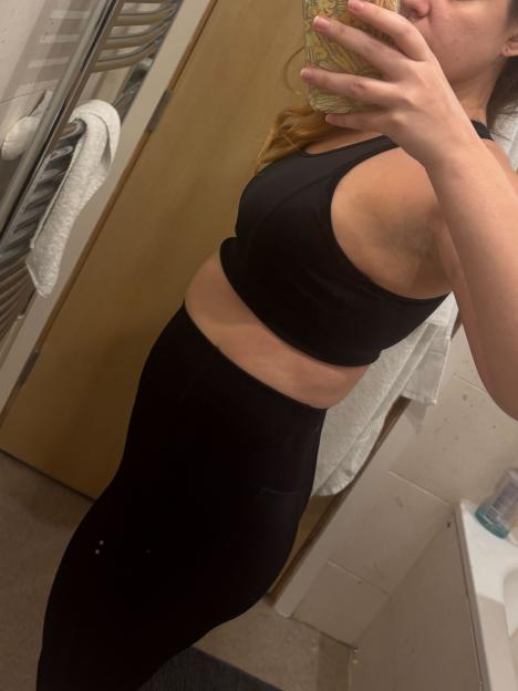 A woman in black sports bra and leggings, viewed from the side, shows her midsection.