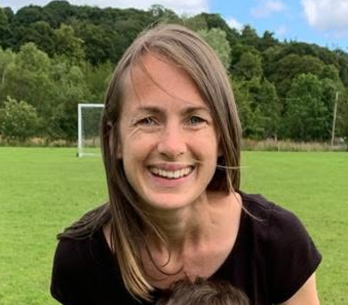 Elisabeth-Jane Ross smiling in an outdoor setting with a soccer goal and trees in the background.