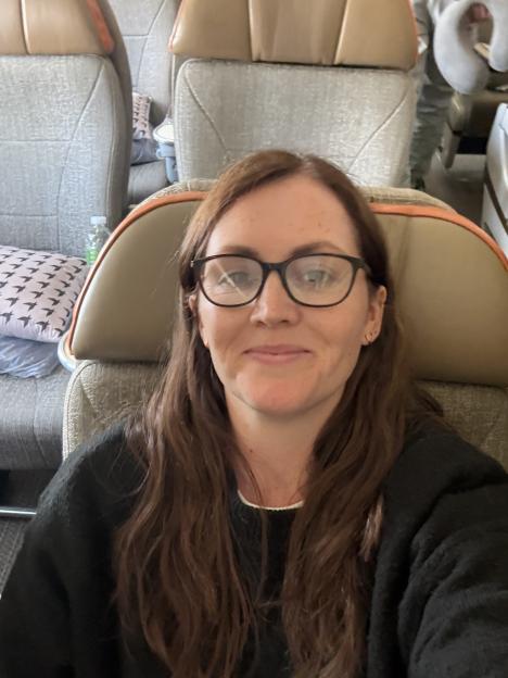 A woman with long brown hair and glasses sitting in an airplane seat.
