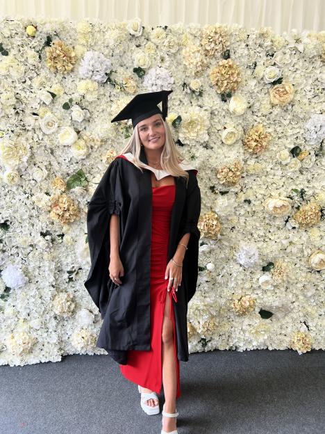 Emily Rose Browning in her graduation attire, consisting of a black cap and gown over a red dress, standing against a floral backdrop.