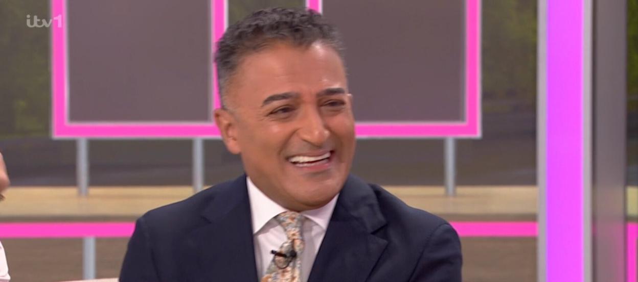 A man in a suit jacket, white shirt, and patterned tie laughs on the set of Good Morning Britain, with ITV channel logos and a news banner at the bottom.