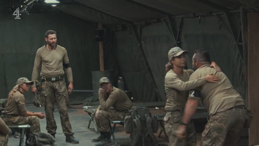 Ryan Thomas wearing a tan long-sleeved shirt and camouflage pants, looking distressed with other people in military-style uniforms in a tent.