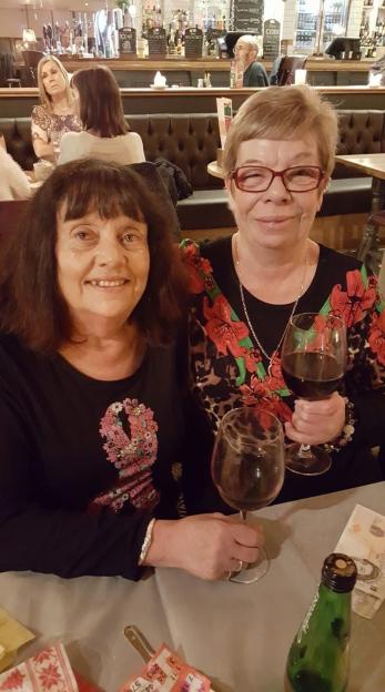Two women smiling, holding glasses of red wine, with other patrons visible in the background of a restaurant.