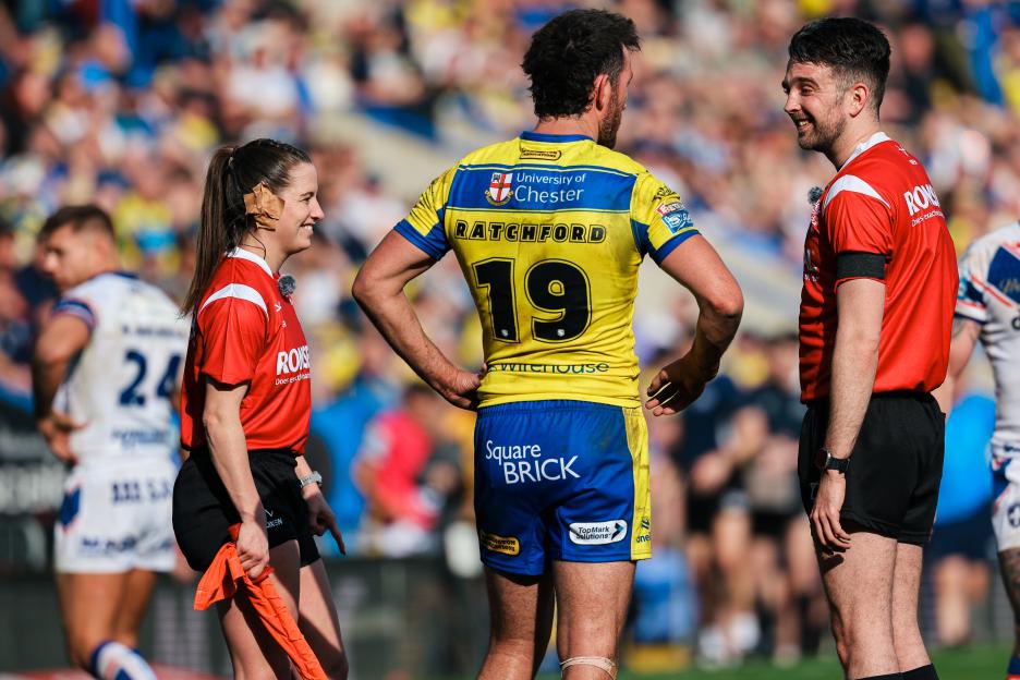 Tara Jones, the Super League touch judge, smiles on a rugby field alongside two male players.