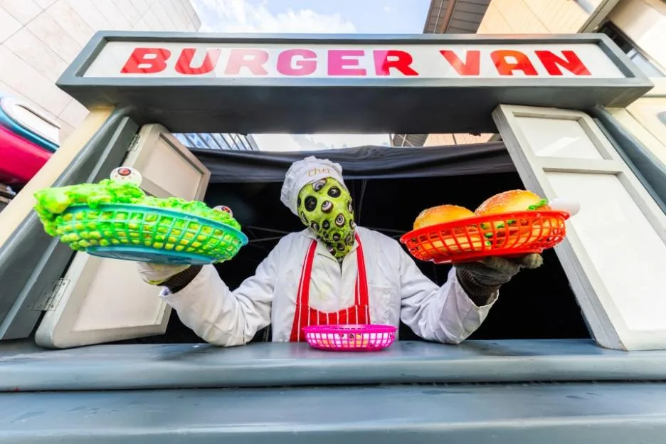 A person wearing a chef's uniform and a mask covered in eyeballs stands inside a "Burger Van," holding out two baskets of food.