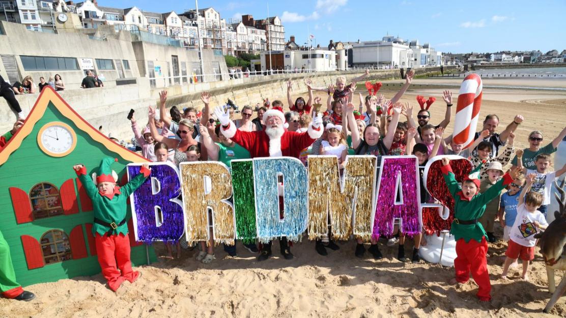 A crowd of people, including children dressed as elves and Santa Claus, pose on a beach behind large, sparkly letters spelling "BRIDMAS."