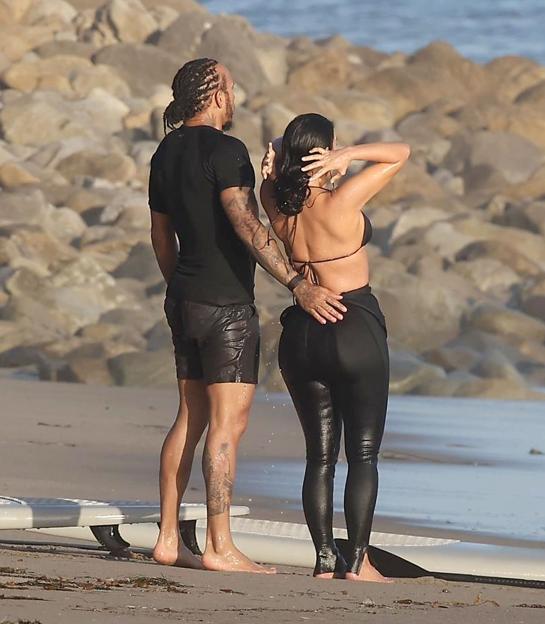 Kim Kardashian in a black bikini top and black leggings and Lewis Hamilton in a black t-shirt and black shorts on a beach with a surfboard.