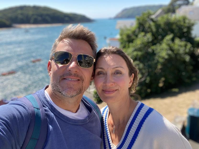 Ben Shephard and his wife in front of a bay with green hills.