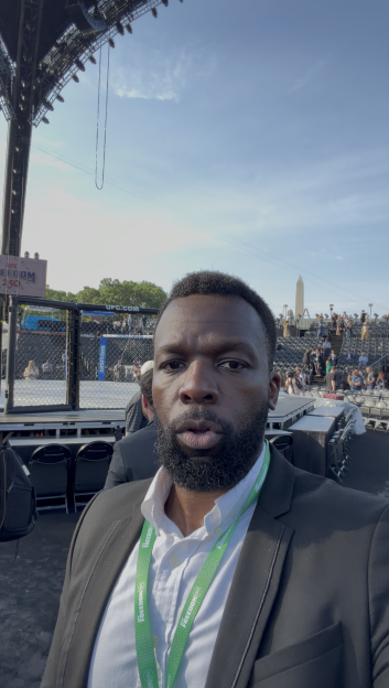 A man in a suit jacket and white shirt, wearing a green lanyard, takes a selfie in front of a UFC octagon, with empty seating and the Washington Monument in the background.